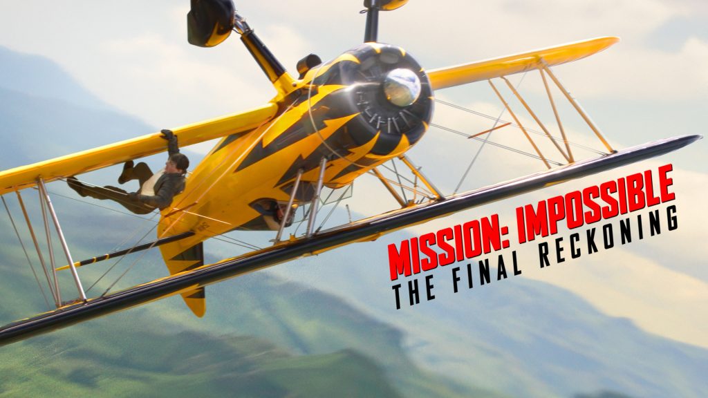 Tom Cruise berpegangan pada sayap pesawat biplane kuning yang sedang terbang terbalik di udara, menampilkan aksi akrobatik berbahaya dengan latar belakang pegunungan hijau, terdapat teks merah "MISSION: IMPOSSIBLE THE FINAL RECKONING" di pojok kanan bawah.