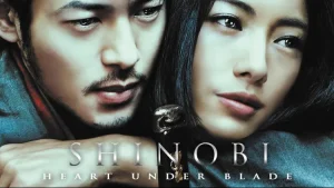 Foto close-up poster film Shinobi: Heart Under Blade menampilkan wajah dua karakter utama; pria di sisi kiri (Gennosuke) dengan kumis dan janggut tipis menatap ke bawah, dan wanita di sisi kanan (Oboro) dengan rambut hitam panjang menatap ke arah depan dengan ekspresi melankolis; terdapat teks judul "SHINOBI HEART UNDER BLADE" di bagian bawah gambar.